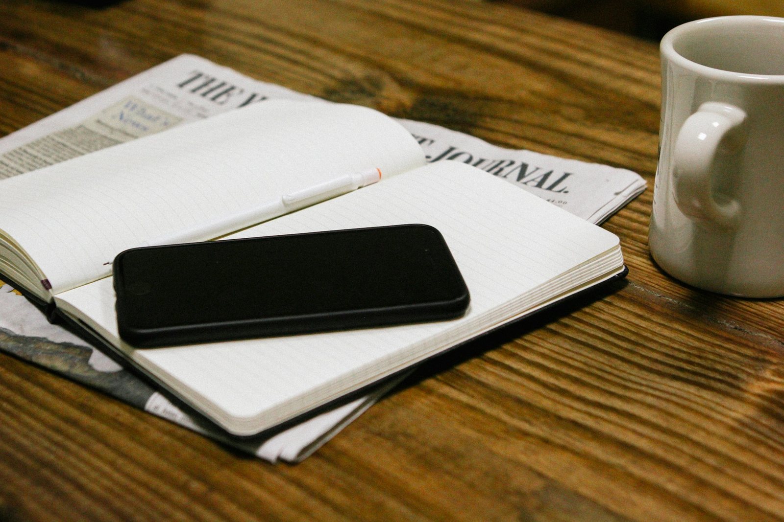 A notebook, newspaper and coffee cup on a wooden desk.